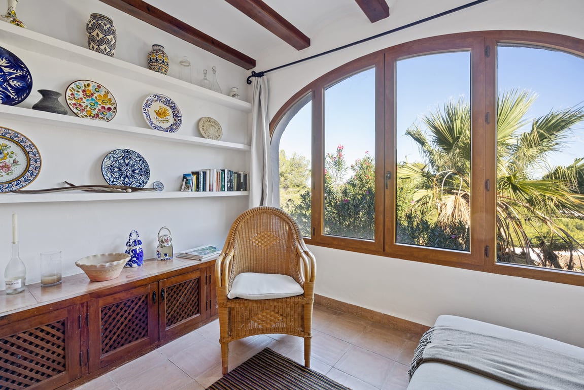 Glazed naya featuring exposed wooden beams, built-in shelving with decorative ceramics, and a large arched window overlooking the Mediterranean garden.