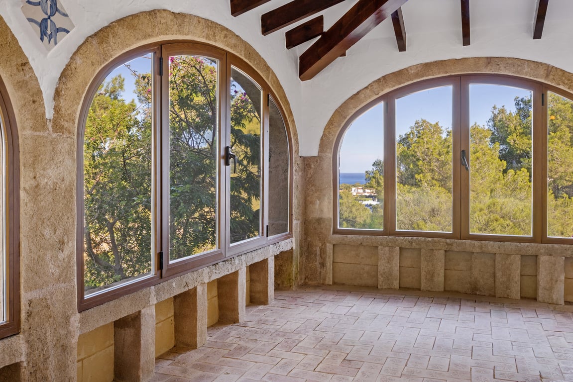 Glazed naya featuring Tosca stone arches, exposed wooden beams, and traditional clay flooring. Large windows provide views of the sea and Mediterranean greenery.