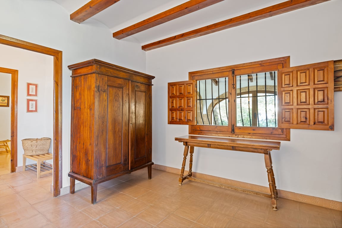 Hallway featuring exposed wooden ceiling beams, traditional ceramic floor tiles, and antique-style furniture. A window with classic shutters overlooks a stone arch.