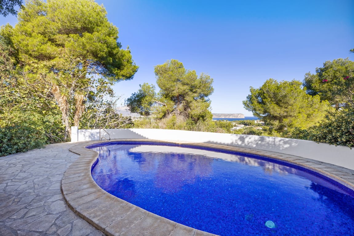 Kidney-shaped private pool featuring blue mosaic tiles and a natural stone terrace, offering distant sea views through mature Mediterranean pines.