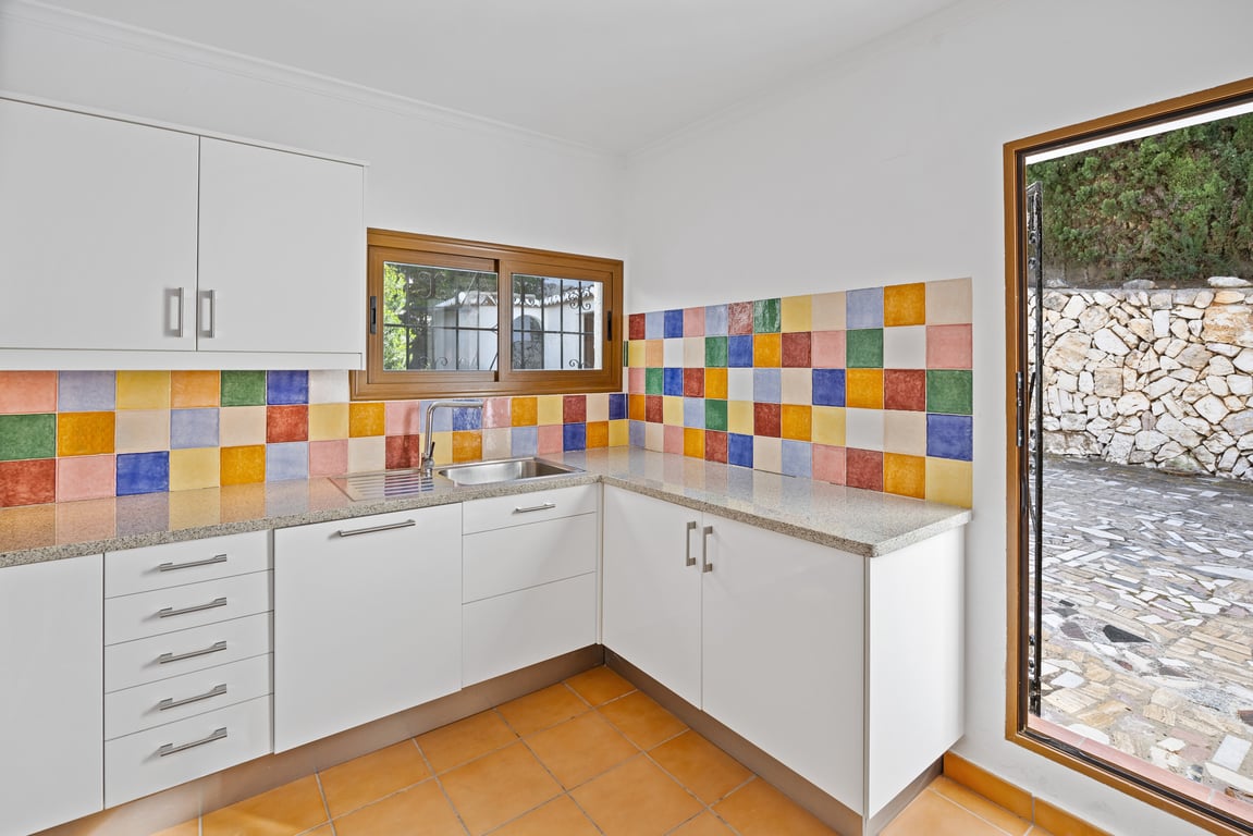 Kitchen featuring white cabinetry, granite countertops, and a vibrant multi-colored tiled backsplash. The space includes terracotta flooring and direct access to the exterior.