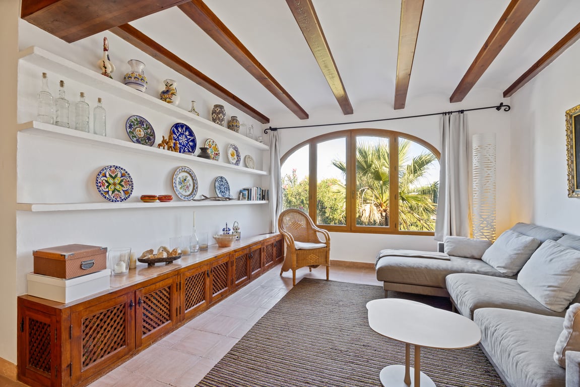 Living area featuring exposed wooden beams, built-in shelving with decorative ceramics, and a large arched window overlooking lush palm trees.