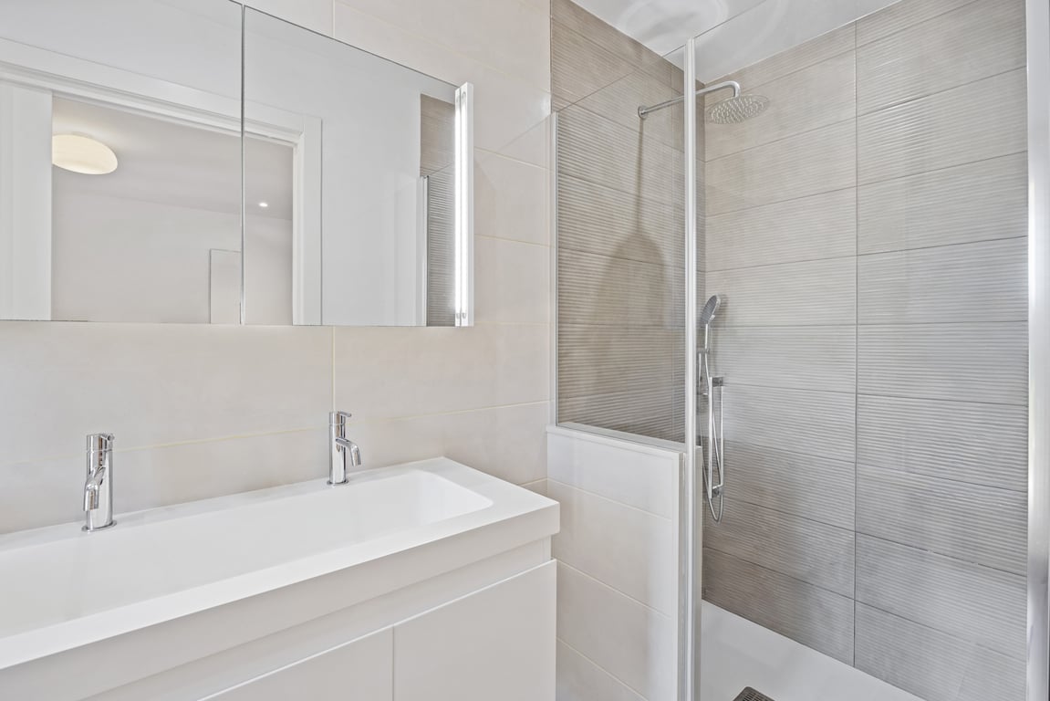 Modern renovated bathroom featuring a double-faucet vanity, large mirrored cabinet, and a walk-in shower with textured grey tiling and glass partition.