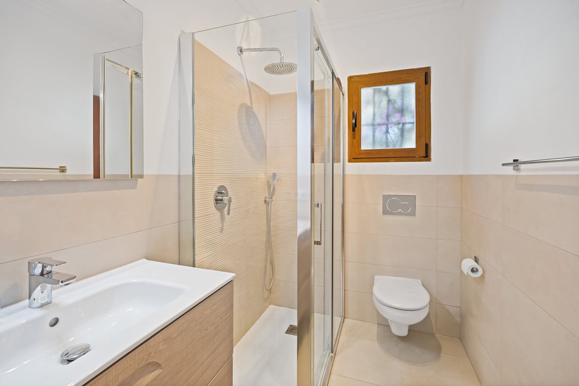 Modern renovated bathroom featuring a walk-in glass shower, light wood vanity with ceramic sink, and neutral beige tiling throughout.
