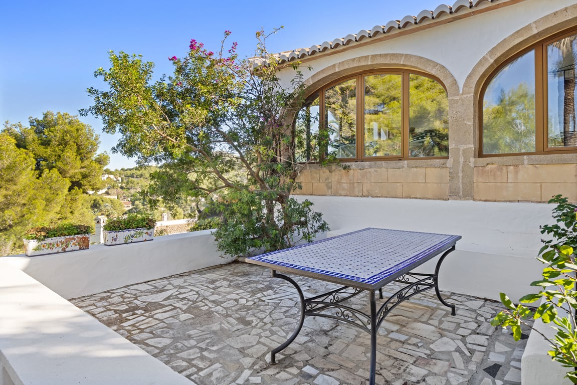 Outdoor terrace featuring irregular stone paving, a traditional tiled table, and Tosca stone arches framing the glazed naya against a backdrop of pine trees.