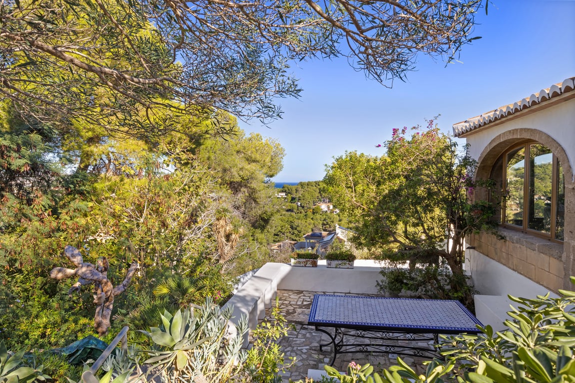 Outdoor terrace featuring natural stone paving and a mosaic tile table, surrounded by Mediterranean greenery with sea views framed by pines and traditional architecture.