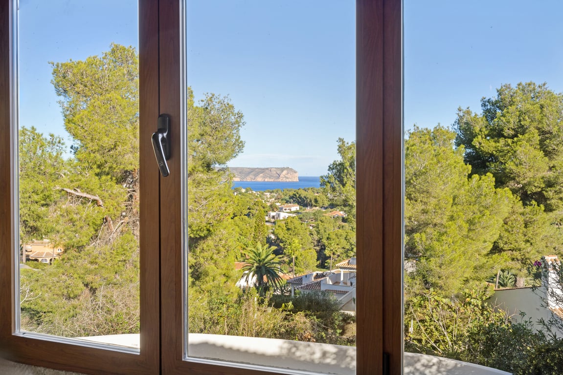 Panoramic sea and headland views framed by a wood-effect window with double glazing, overlooking lush Mediterranean pine trees.