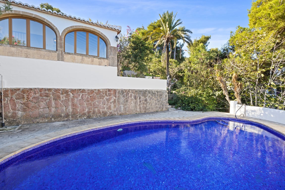 Private swimming pool featuring blue mosaic tiling, bordered by a rustic stone wall and a terrace, with the villa's arched naya and lush palms in the background.