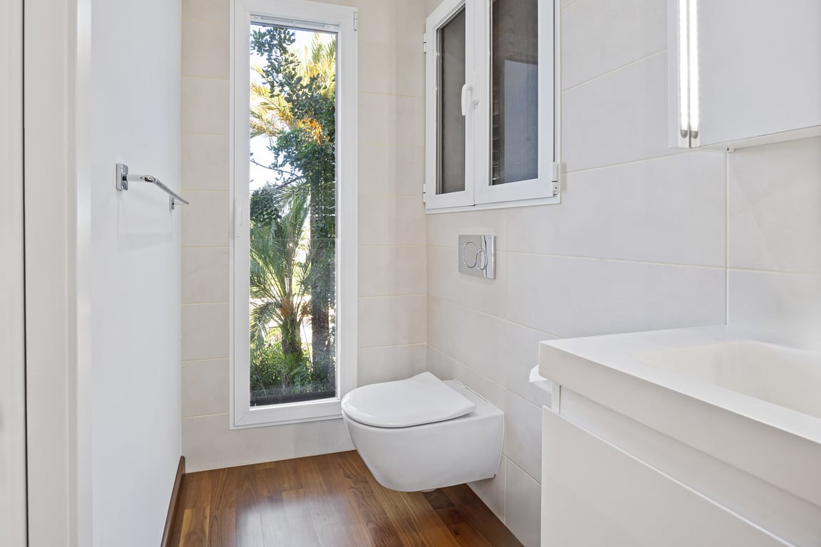 Renovated bathroom featuring a wall-hung toilet, wood-effect flooring, and a vertical window overlooking lush garden greenery.