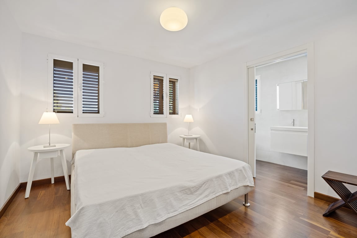Renovated bedroom featuring dark wood flooring, a fabric headboard, and a sliding door leading to a modern en-suite bathroom with a floating vanity.