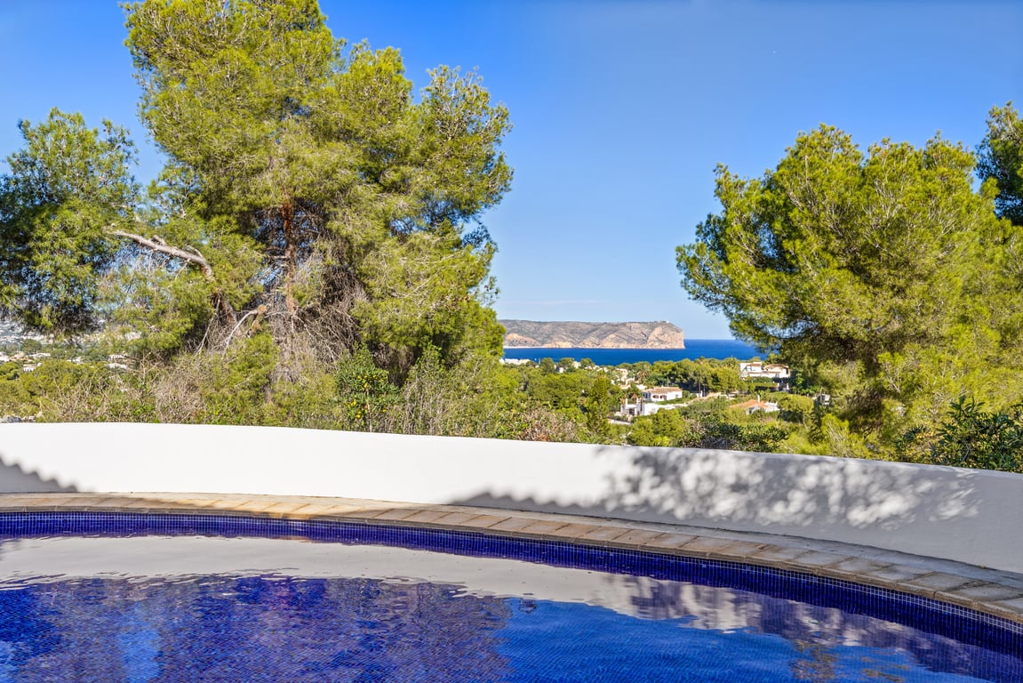 View from the private pool overlooking the Mediterranean Sea and coastal cliffs, framed by mature pine trees and a curved white wall.