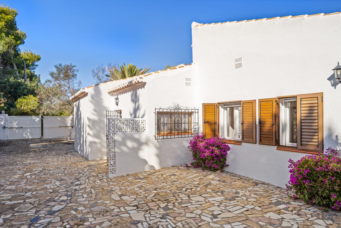 Villa exterior showing the white facade, wooden shutters, wrought iron grilles, and irregular stone paving bordered by purple bougainvillea.