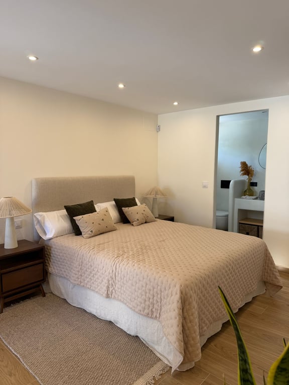 Bright bedroom featuring a double bed with upholstered headboard, wooden nightstands, and recessed lighting. An en-suite bathroom with modern vanity is visible.