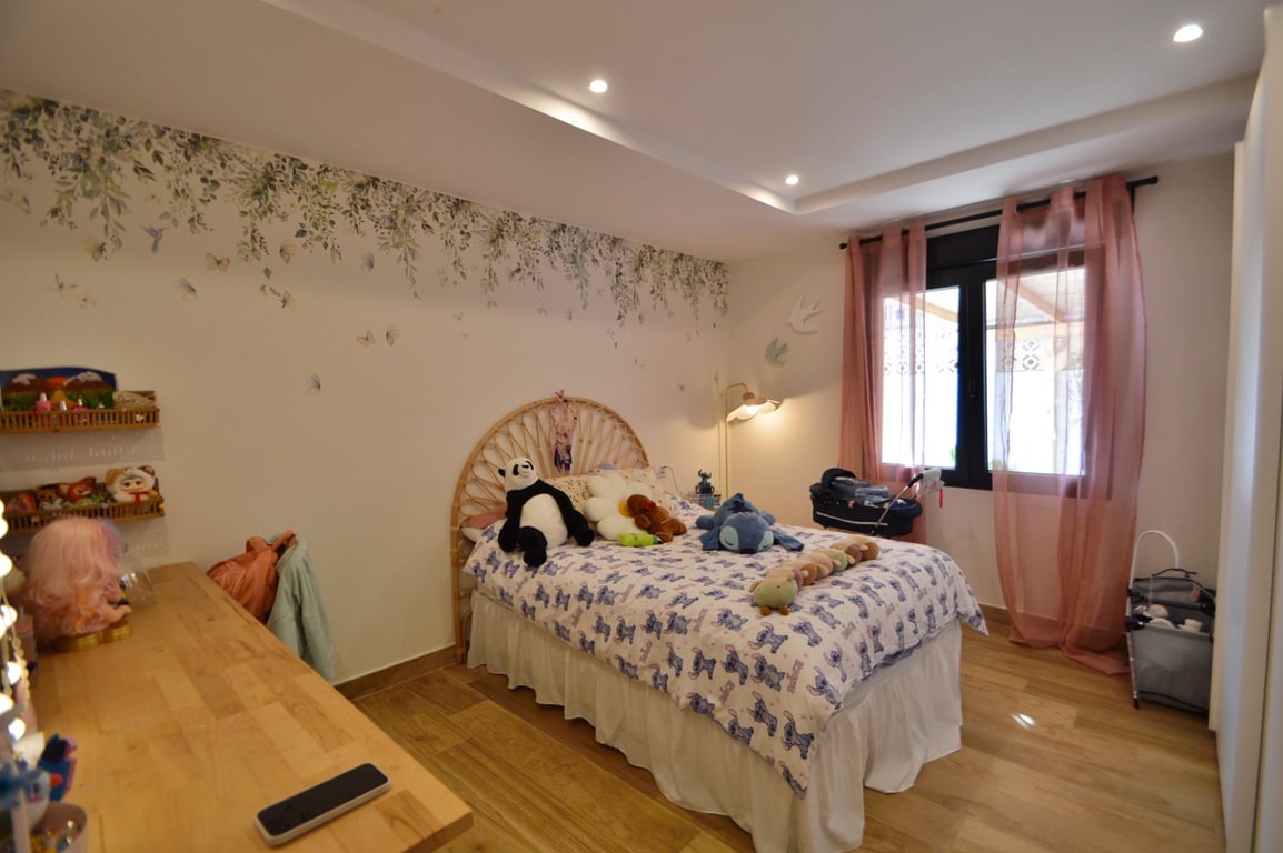 Bright bedroom featuring a rattan headboard, botanical wall mural, and warm wood flooring. The space is finished with recessed ceiling lights and soft natural lighting.