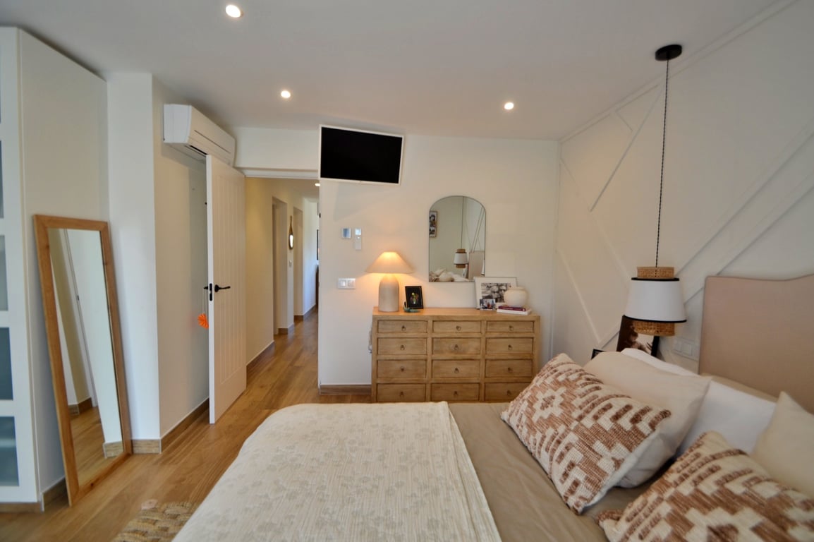 Bright bedroom featuring wood-effect flooring, geometric wall paneling, split air conditioning, and natural wood furniture including a large dresser and full-length mirror.