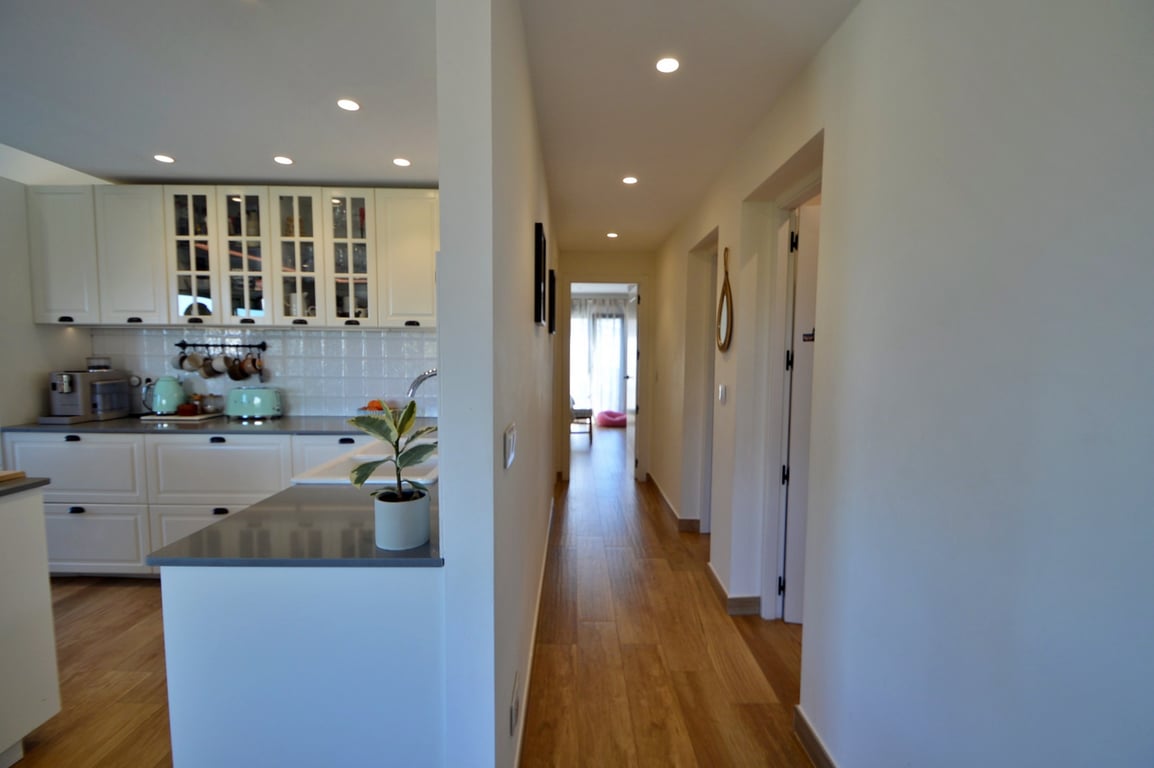 Bright hallway with wood-effect flooring leading past a modern kitchen featuring white cabinetry, glass-fronted cupboards, and recessed ceiling lighting.
