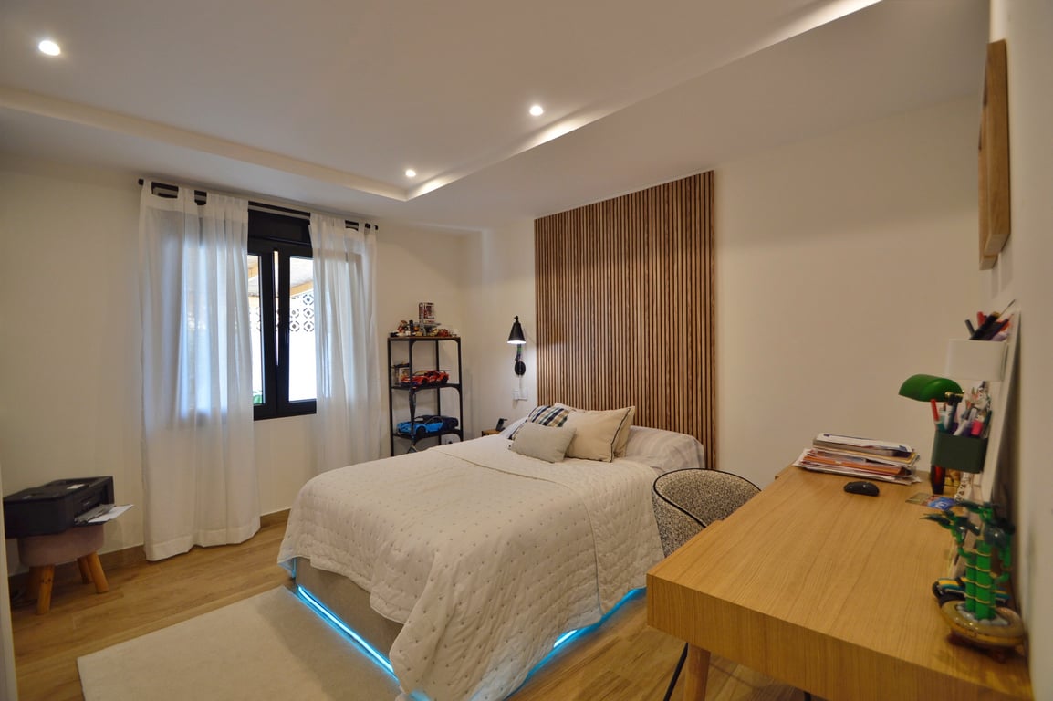 Contemporary bedroom featuring a vertical wood slat headboard, integrated desk, and bed with blue LED accent lighting. Includes wood-effect flooring and recessed ceiling spots.
