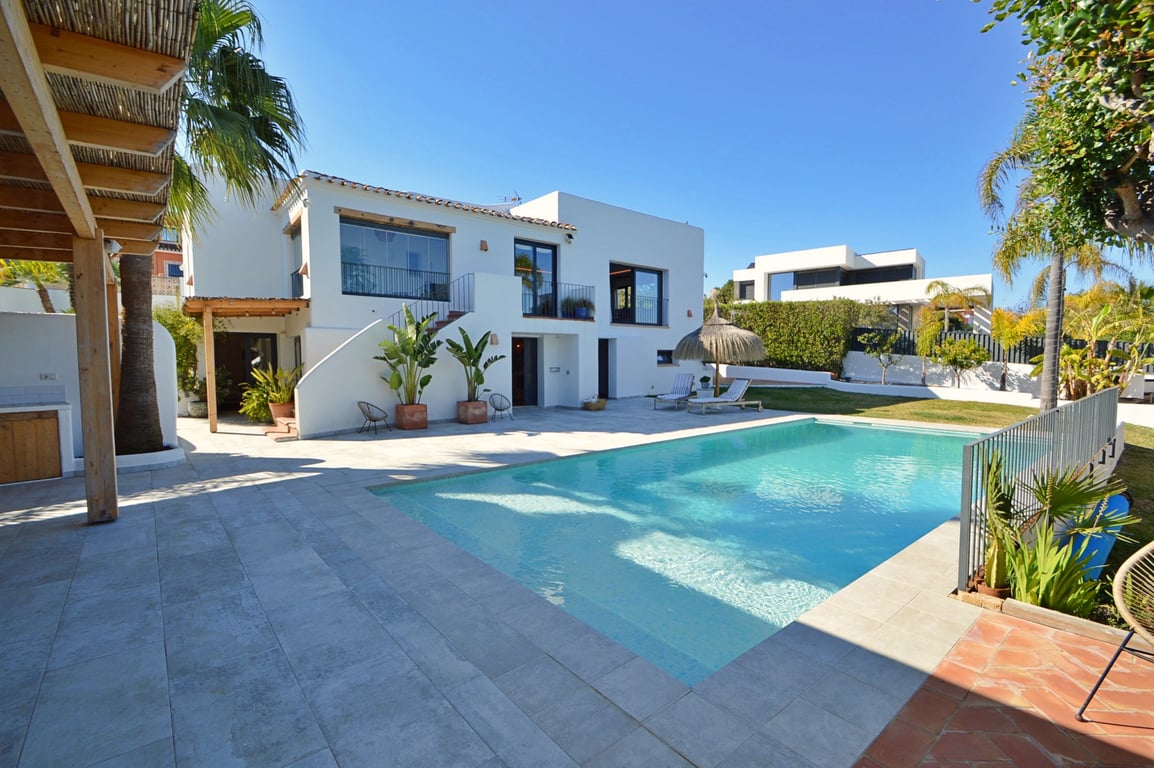 Exterior view of the villa featuring a large swimming pool, light stone terrace, wooden pergola, and a white Ibiza-style facade with a glass-enclosed upper naya.