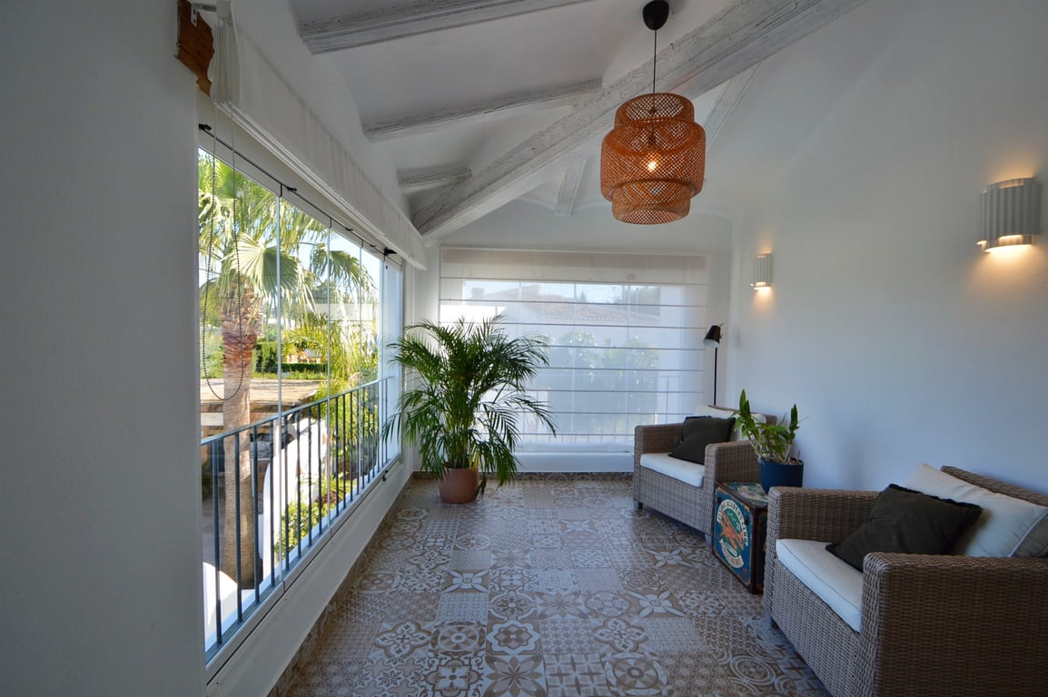 Glass-enclosed naya featuring white-painted wooden beams, patterned ceramic tiles, and wicker seating with views of the lush garden.