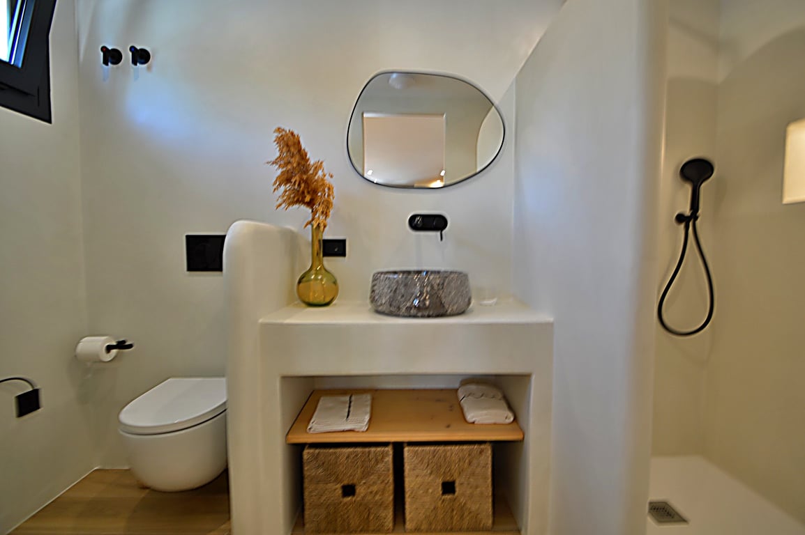 Ibiza-style bathroom featuring microcement walls, a stone vessel sink, and matte black fixtures. An organic-shaped mirror and built-in vanity with wicker storage complete the space.