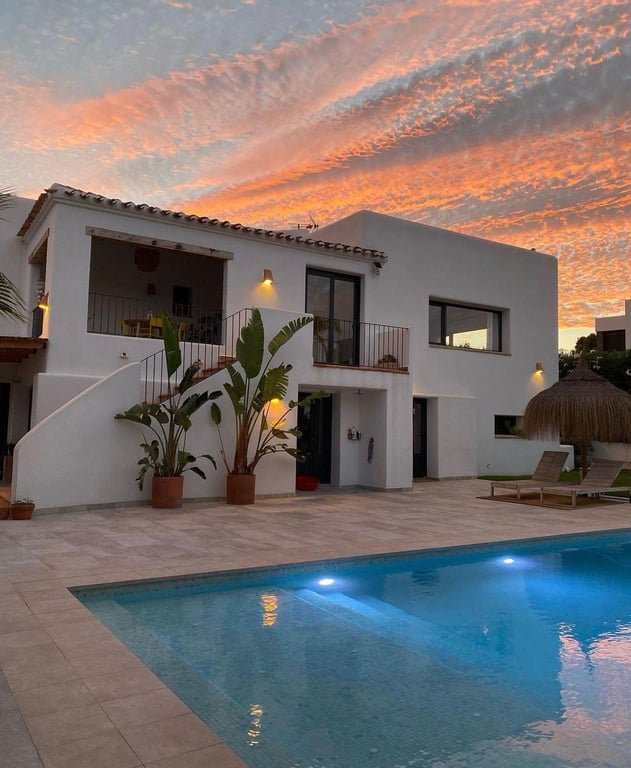 Ibiza-style villa exterior at dusk, showcasing the illuminated saltwater pool, natural stone terrace, and Mediterranean landscaping against a sunset sky.
