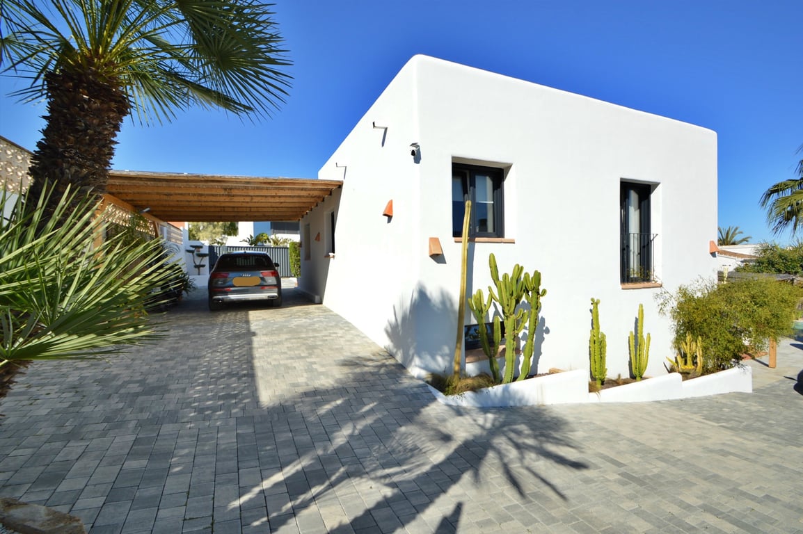 Ibiza-style villa exterior featuring a wooden beam carport, paved driveway, and Mediterranean landscaping with cacti and palm trees.