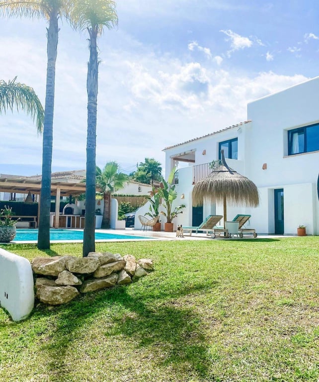 Ibizan-style exterior featuring a manicured lawn, swimming pool, and thatched umbrella. The background reveals the outdoor kitchen with a pizza oven.