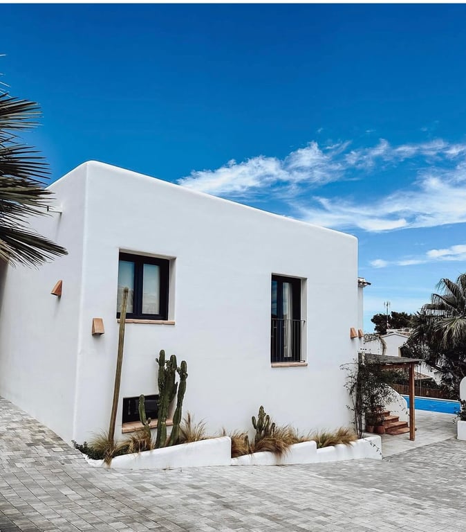 Ibizan-style exterior featuring smooth white walls, dark-framed windows, and a minimalist cactus garden alongside a grey cobblestone driveway.