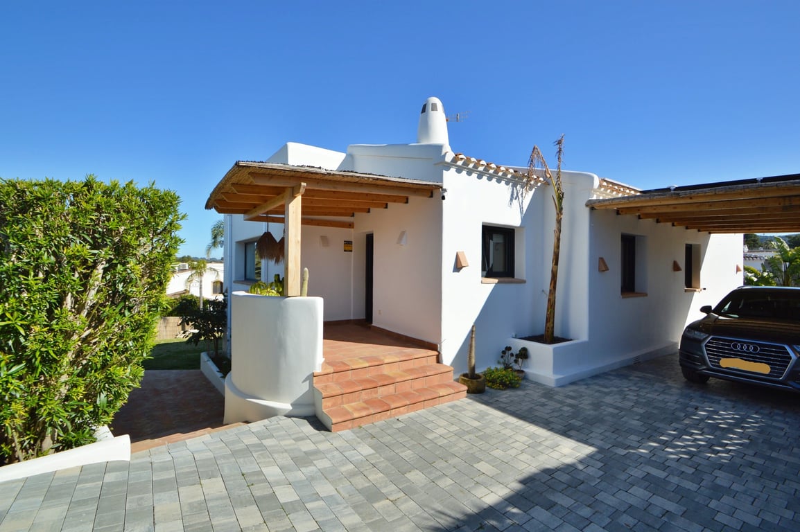 Ibizan-style main entrance featuring a wooden pergola with reed roofing, terracotta steps, and a paved driveway leading to a covered carport.