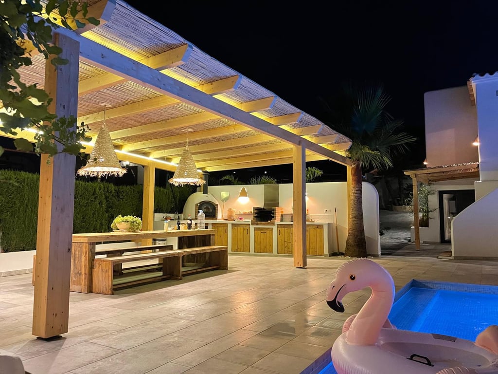 Illuminated outdoor kitchen featuring a wood-fired oven, BBQ, and dining area under a timber pergola with integrated LED lighting.