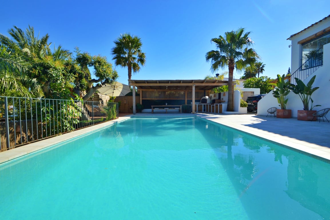 Large rectangular swimming pool with a spacious sun terrace, featuring a covered wooden pergola for outdoor dining and lush Mediterranean landscaping.