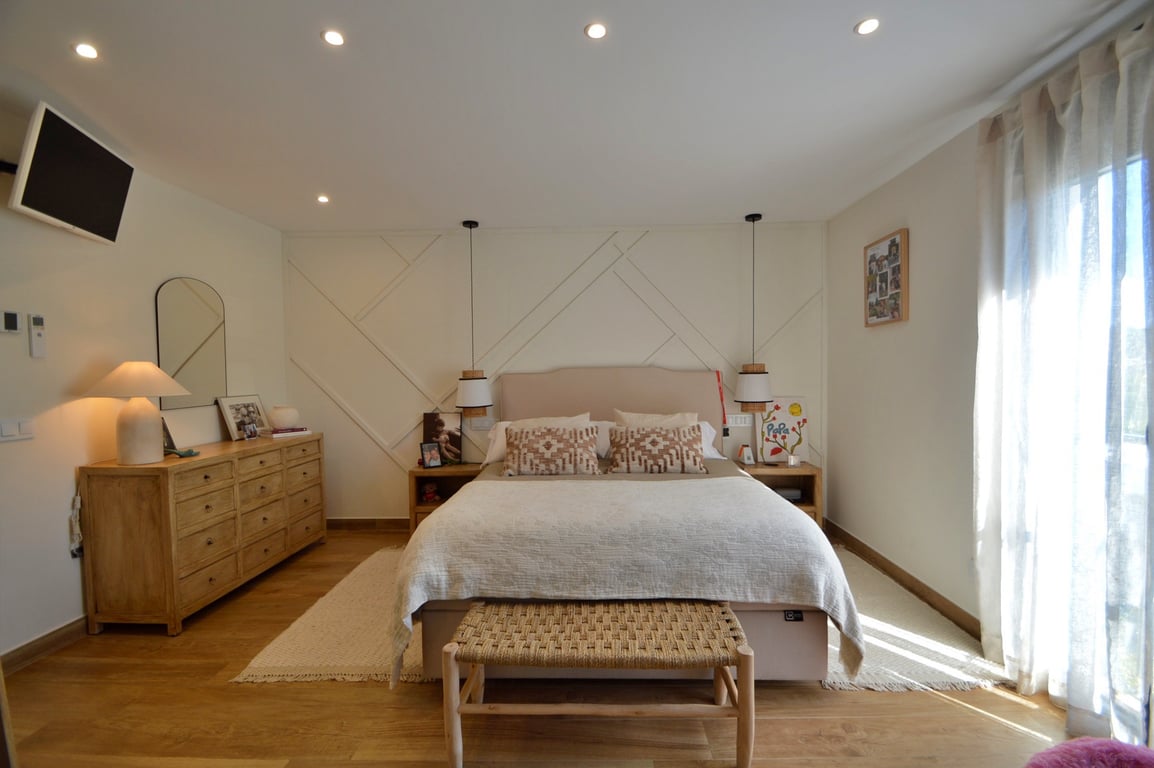 Master bedroom featuring a geometric accent wall, light wood flooring, and integrated lighting. The space includes a wooden dresser, woven bench, and large window with sheer curtains.