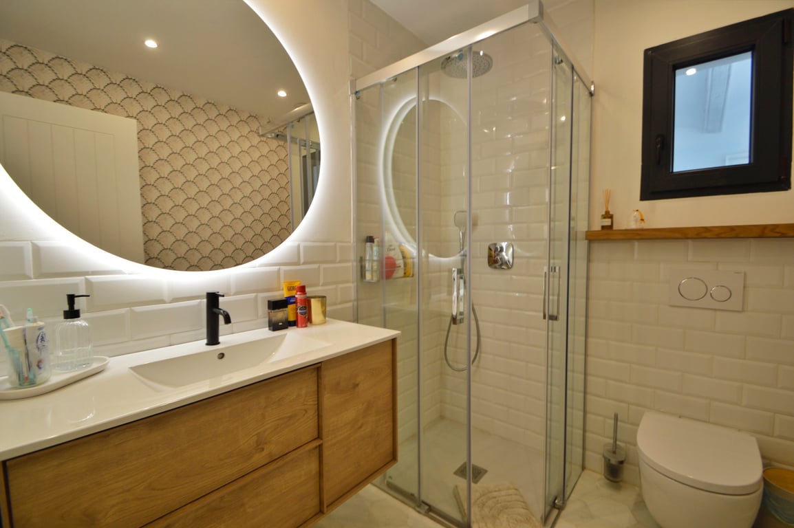 Modern bathroom featuring a backlit circular mirror, wooden vanity with black fixtures, and a glass shower with white subway tiles.
