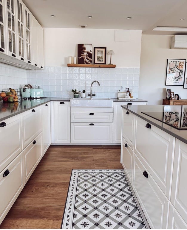 Modern country-style kitchen featuring white cabinetry, dark countertops, and a farmhouse sink. The design includes a wooden floor with a distinctive hydraulic tile inlay.