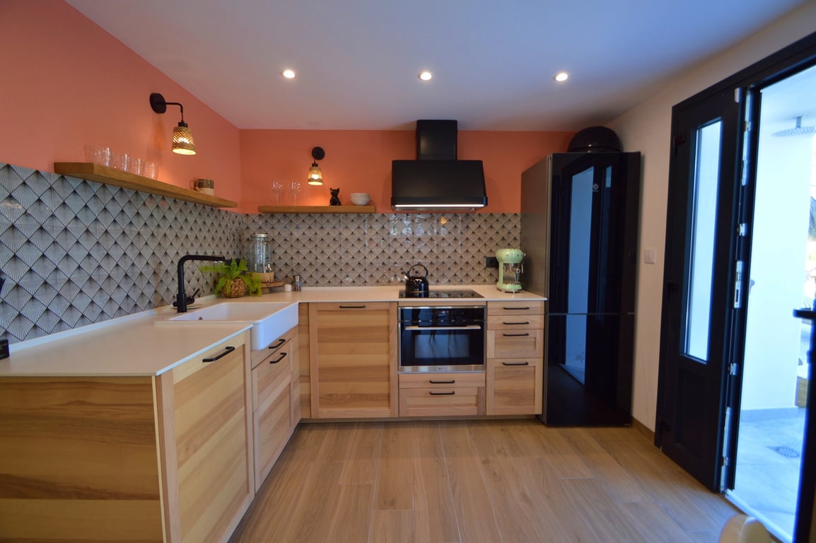 Modern kitchen featuring light wood cabinetry, a white farmhouse sink, and a geometric tile backsplash. Black appliances contrast with the terracotta accent wall and recessed lighting.
