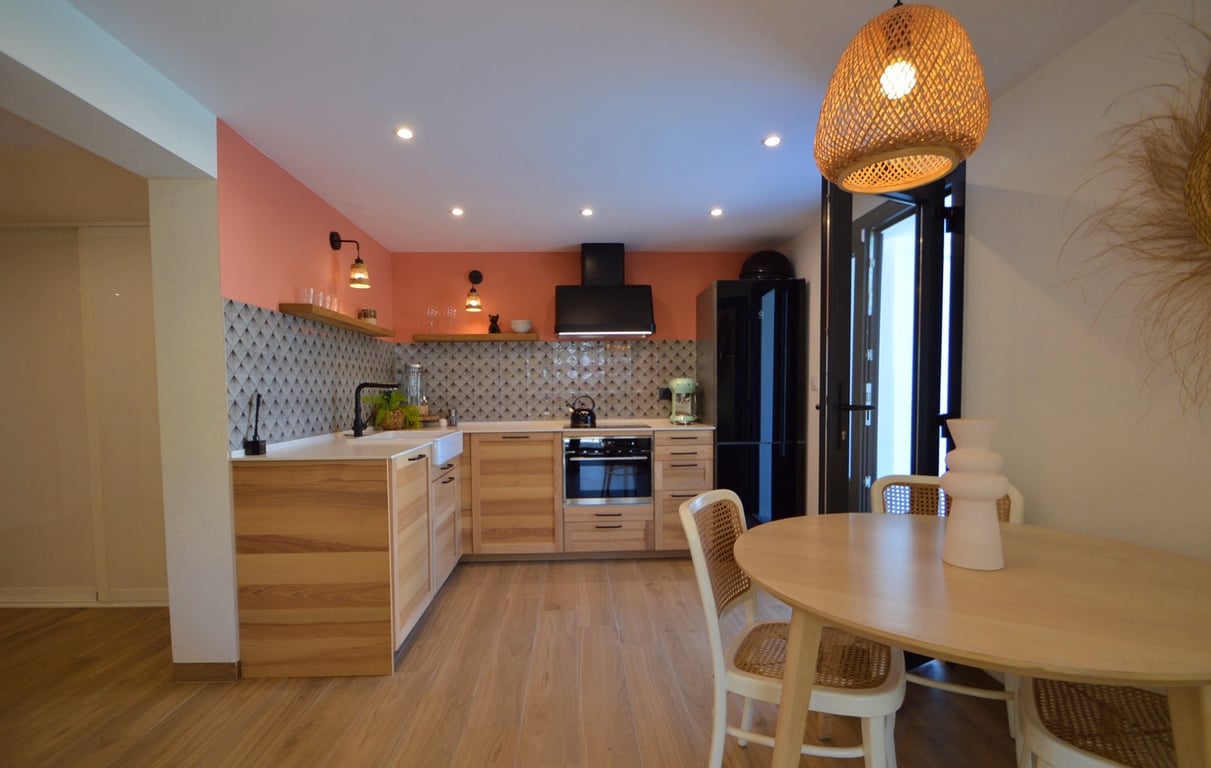 Modern kitchen featuring wooden cabinetry, a terracotta accent wall, and a dining area with a round table, rattan-backed chairs, and a woven fiber pendant light.