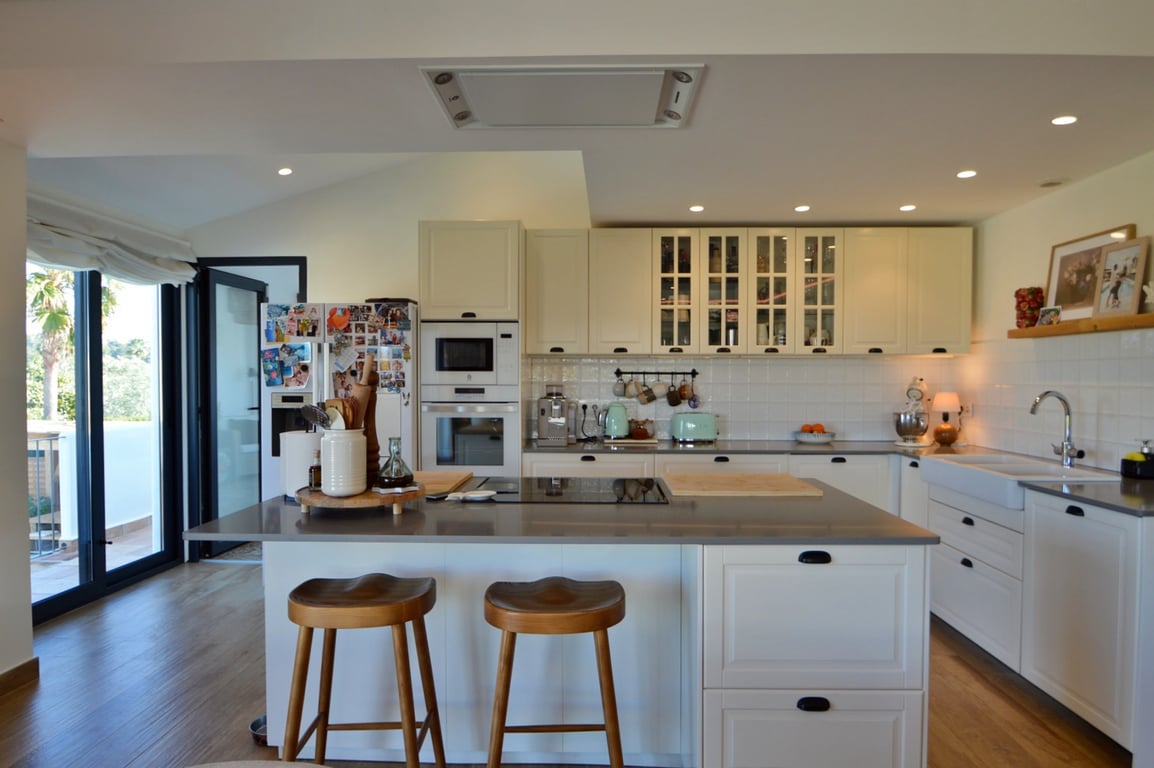 Modern open-concept kitchen featuring a central island with wooden stools, integrated appliances, and white cabinetry. Large glass doors provide abundant natural light.