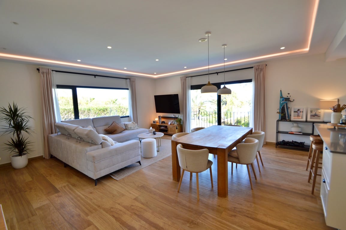 Open-concept living and dining area featuring integrated LED perimeter lighting, warm wooden flooring, and large windows offering views of the garden.