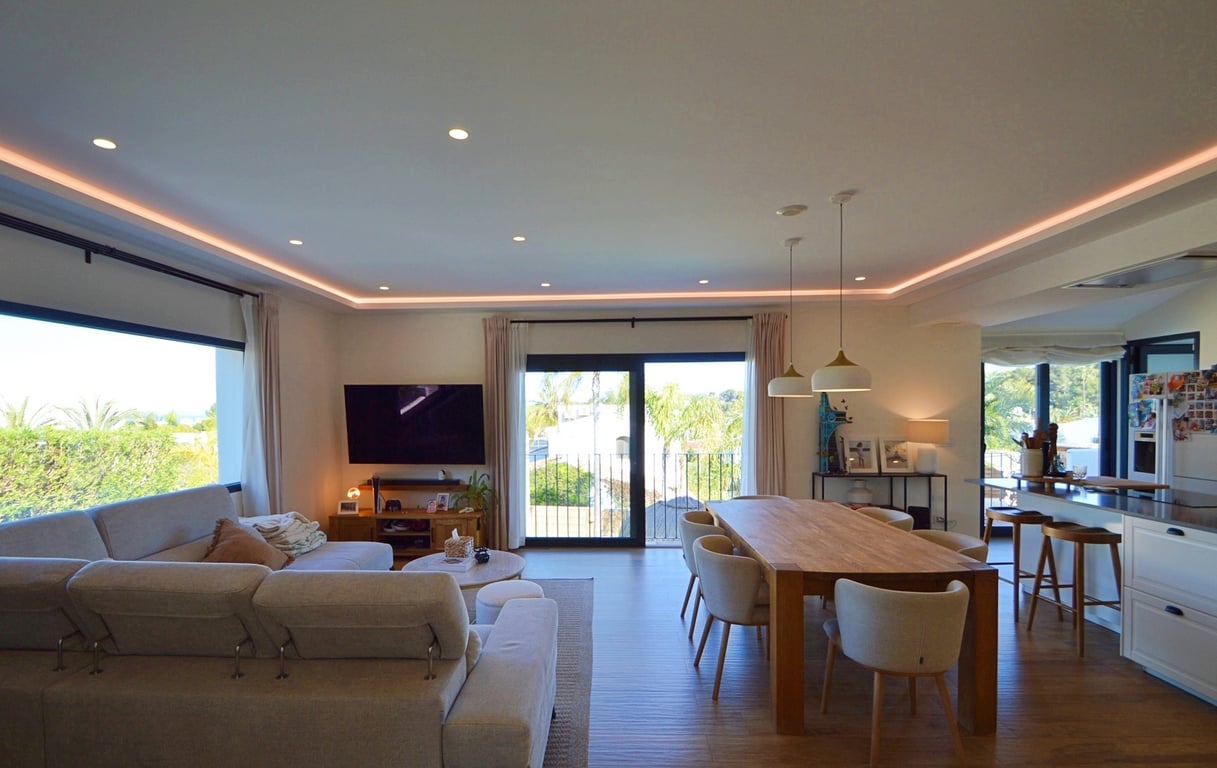 Open-concept living and dining area featuring perimeter LED lighting, a large wooden table, and expansive windows offering views of the surrounding greenery.