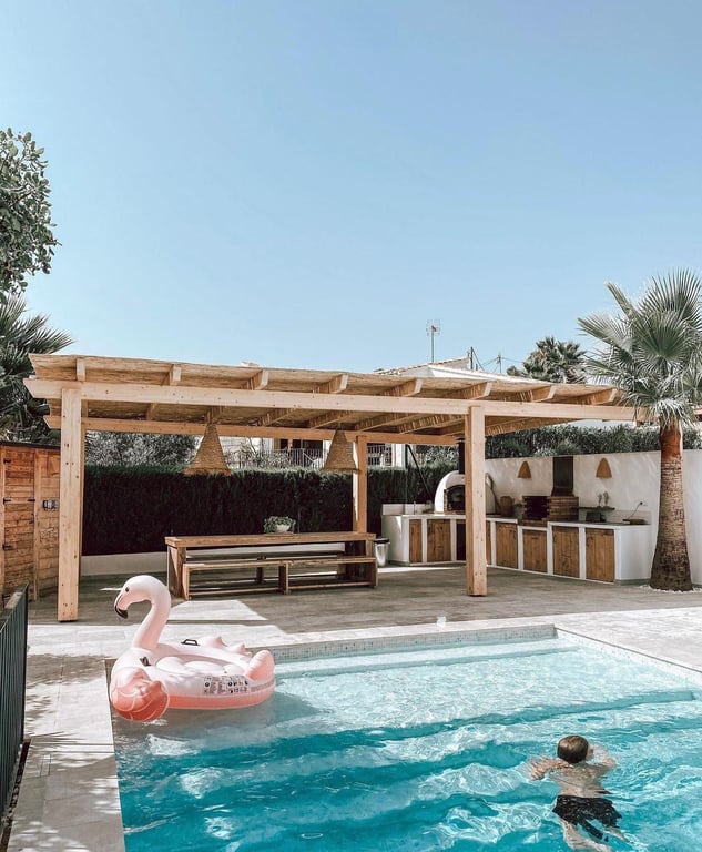 Outdoor dining area and summer kitchen with a pizza oven and barbecue, sheltered by a wooden pergola next to the salt water swimming pool.