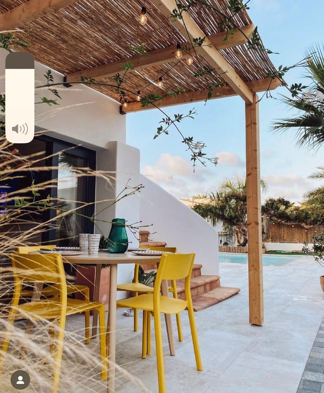 Outdoor dining area featuring a wooden pergola with reed roofing and string lights. Bright yellow chairs sit on a stone terrace overlooking the pool.