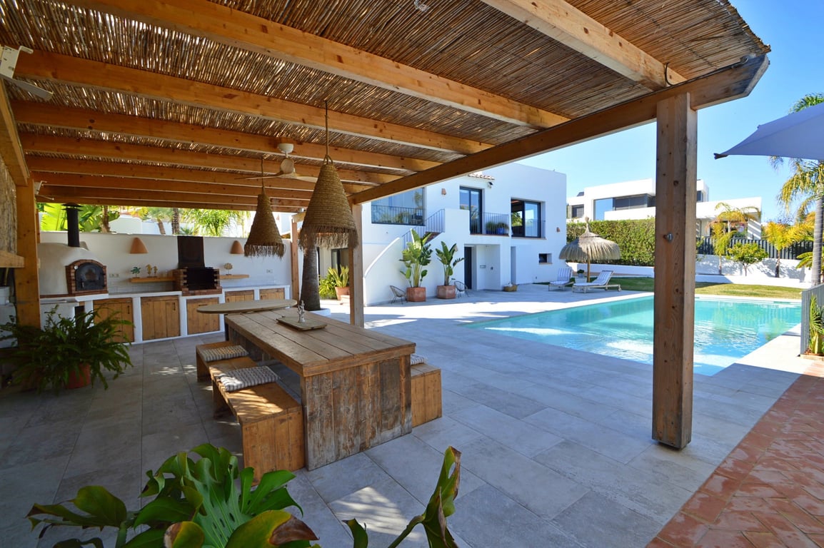 Outdoor kitchen under a timber pergola featuring a pizza oven, rustic dining area, and views of the salt-water pool and Mediterranean villa.