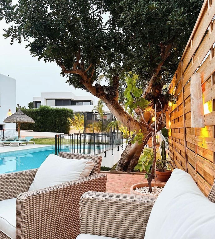 Outdoor lounge area featuring wicker seating with white cushions, terracotta tiling, and a wooden slat fence with warm lighting overlooking the swimming pool.