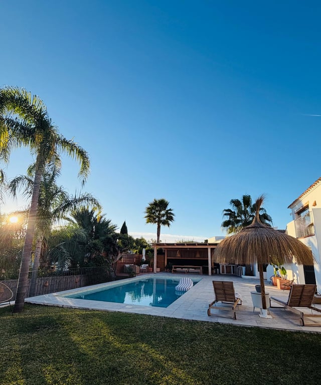Outdoor pool area featuring a stone terrace, heather parasol, and wooden loungers. A covered outdoor kitchen and dining space sit adjacent to the landscaped Mediterranean garden.