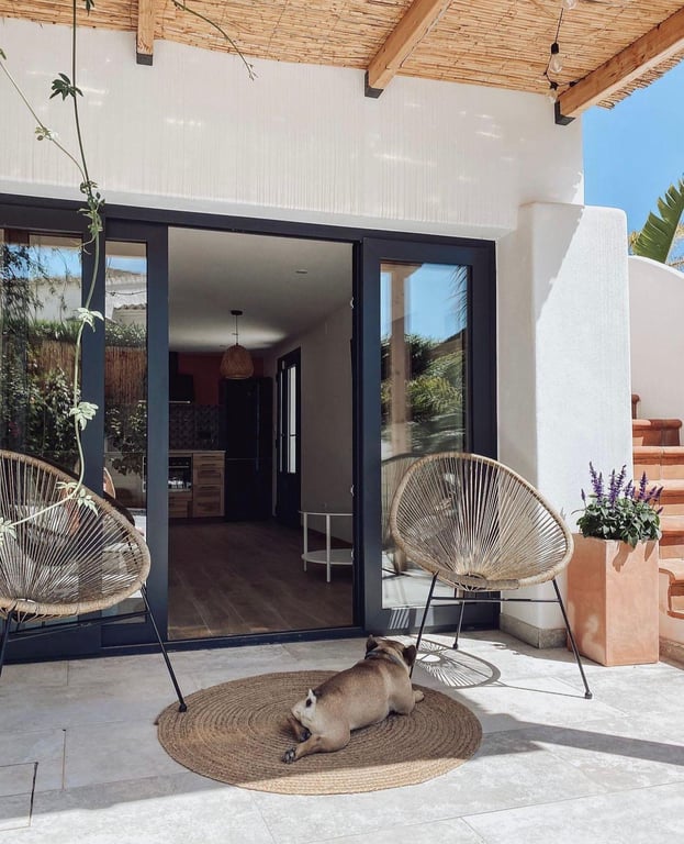 Shaded terrace featuring a natural reed ceiling with wooden beams, dark-framed sliding doors, and contemporary corded seating on stone tiling.