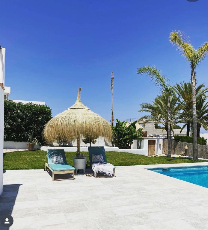 Sun-drenched terrace with teal loungers and a straw parasol beside the saltwater pool, bordered by a green lawn and Mediterranean palm trees.