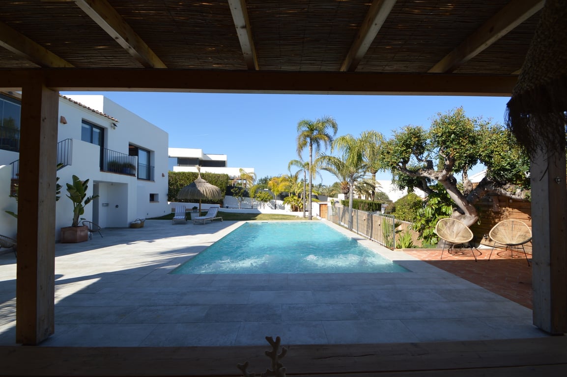 View from the wooden pergola overlooking the saltwater pool, framed by light stone tiling, Mediterranean landscaping, and the white Ibizan-style facade.