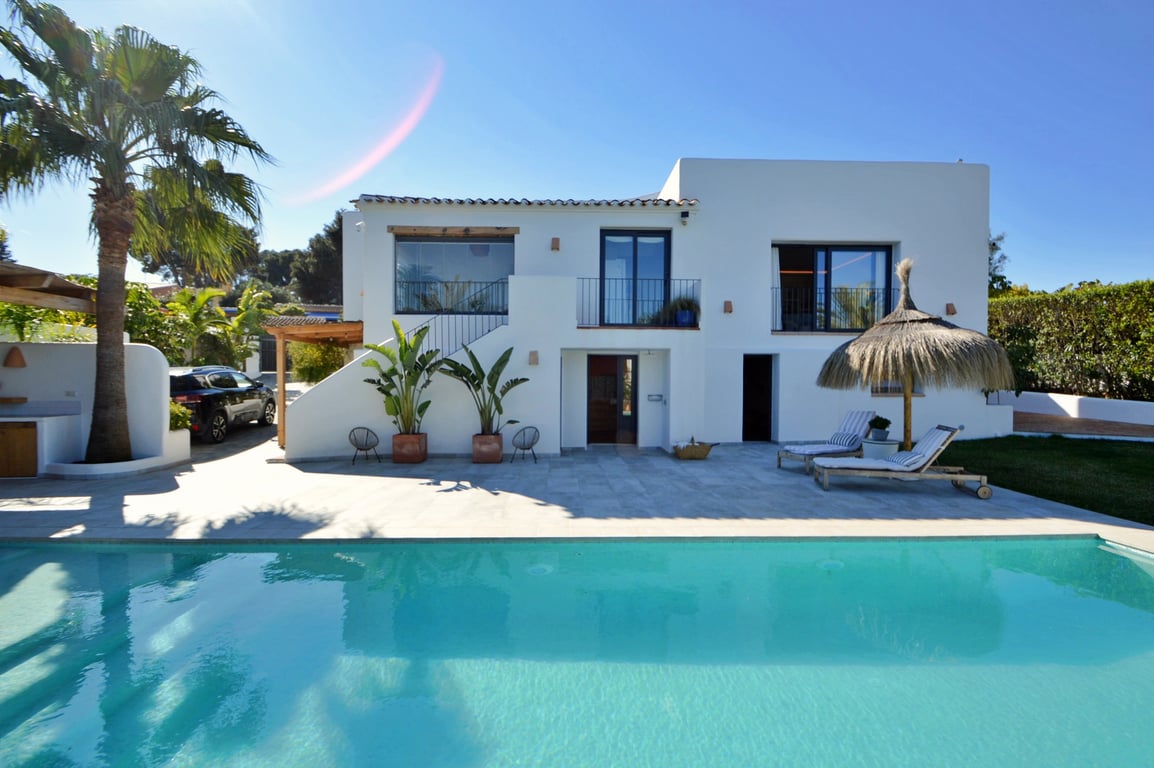 White Ibiza-style facade overlooking a large saltwater pool, featuring a stone terrace, palm trees, and a lounge area with a traditional straw umbrella.