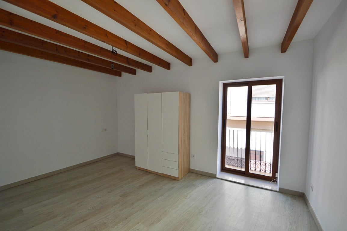 Bedroom featuring exposed wooden ceiling beams, light laminate flooring, and a modern wardrobe. A sliding glass door opens onto a private balcony with iron railings.