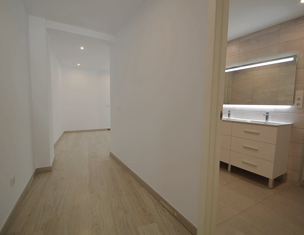 Hallway with light wood flooring leading to a modern bathroom featuring a double vanity, integrated LED mirror, and neutral-toned wall tiles.